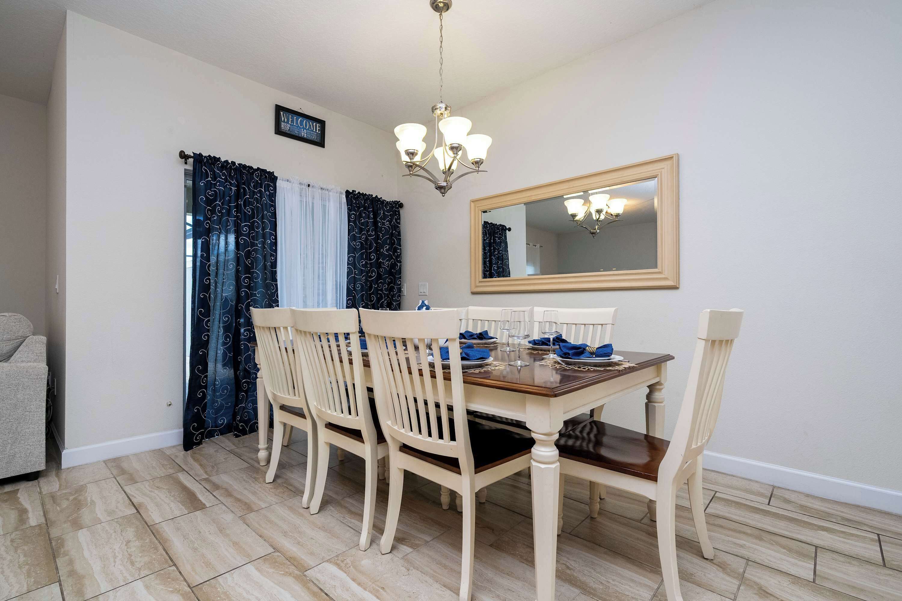 A dining area with a wooden table, four chairs, a chandelier, a mirror, and a sign above the door.