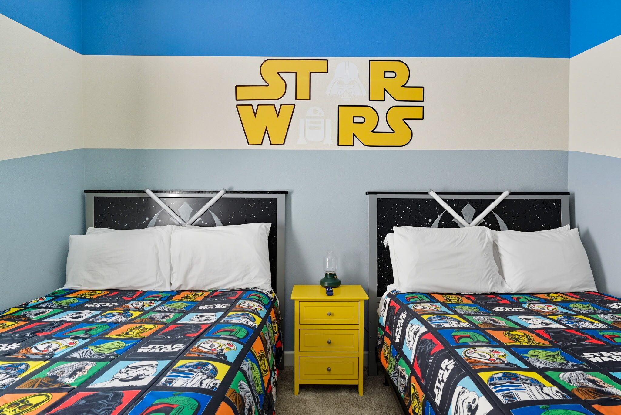 Ein Schlafzimmer mit zwei Betten, beide mit Star Wars Bettwäsche. Dazwischen steht ein gelber Nachttisch, und an der Wand über den Betten steht der Schriftzug 'STAR WARS' in Gelb.