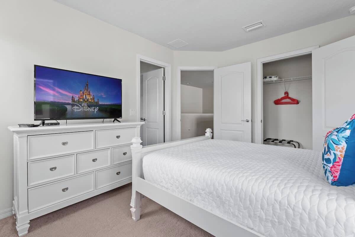 Une chambre avec un lit blanc, une commode, un téléviseur affichant le logo Disney et un placard ouvert avec un cintre rouge.