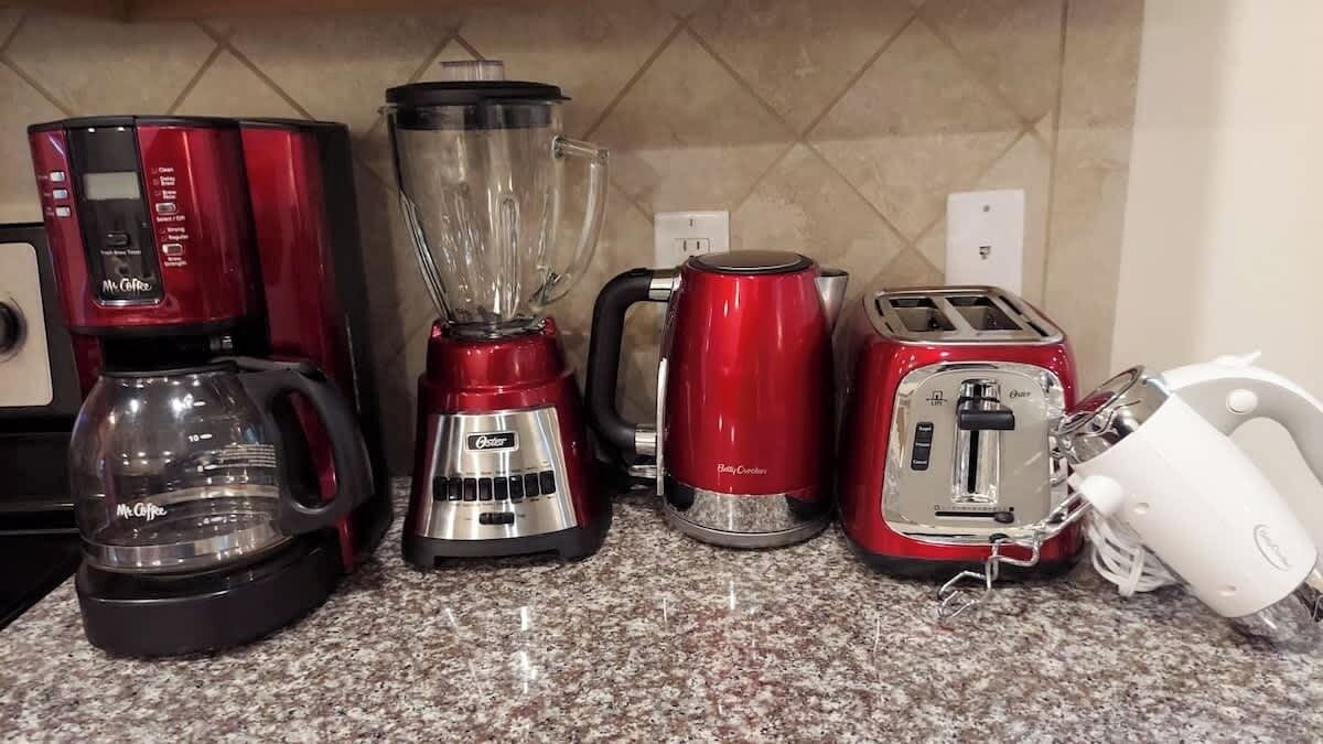 Una encimera de cocina con cafetera, licuadora, hervidor, tostadora y un electrodoméstico blanco, posiblemente un procesador de alimentos.