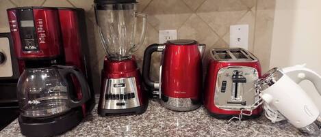 Una encimera de cocina con cafetera, licuadora, hervidor, tostadora y un electrodoméstico blanco, posiblemente un procesador de alimentos.