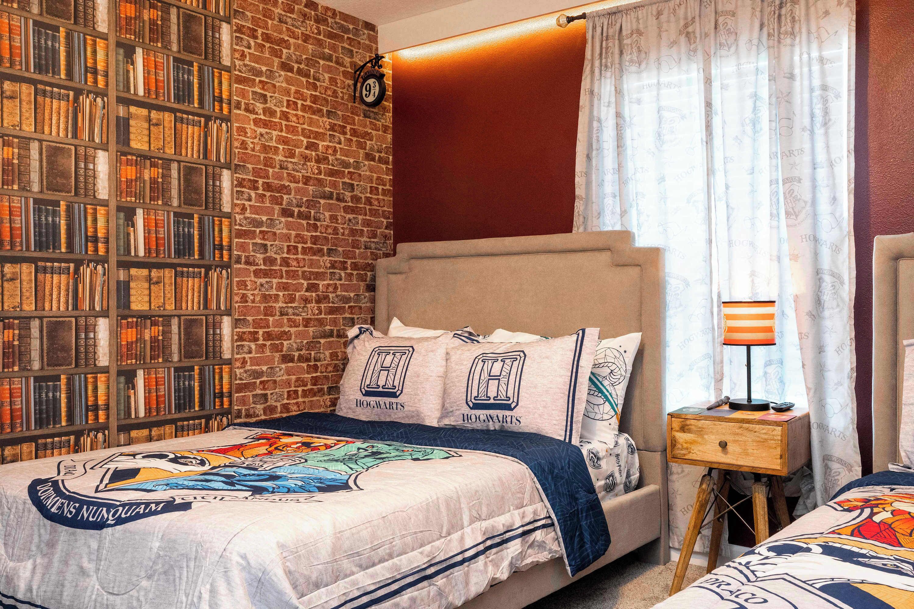 Un dormitorio con pared de ladrillo, estantería, cama con cubrecama de tema Harry Potter y mesita de noche con lámpara.