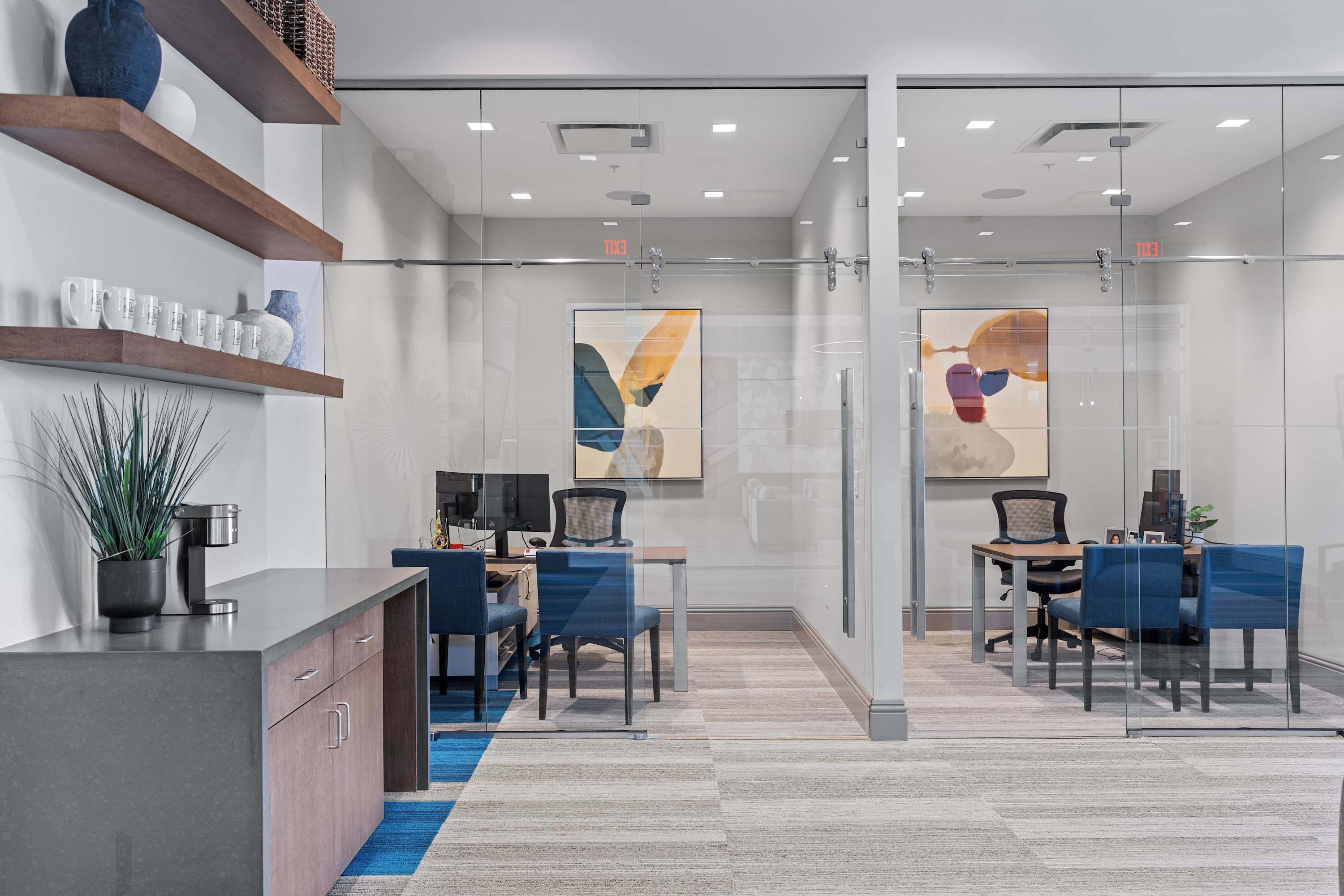 A modern office space with glass partitions, a desk with a computer, chairs, and decorative items on shelves.