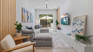 A modern living room with a grey sectional sofa, a wooden side table, and a white armchair. There are two large windows with curtains, a ceiling light, and several wall-mounted paintings. - Luxury Paradise in Orlando Best Location (Orlando)