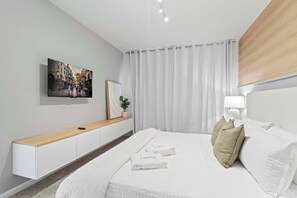 Quarto de hotel moderno com uma cama, televisão de tela plana na parede, mesa de cabeceira e janela com cortinas brancas.