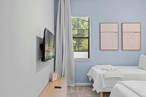 Quarto com televisão, duas obras de arte emolduradas, janela com persianas e cama com roupa de cama branca.