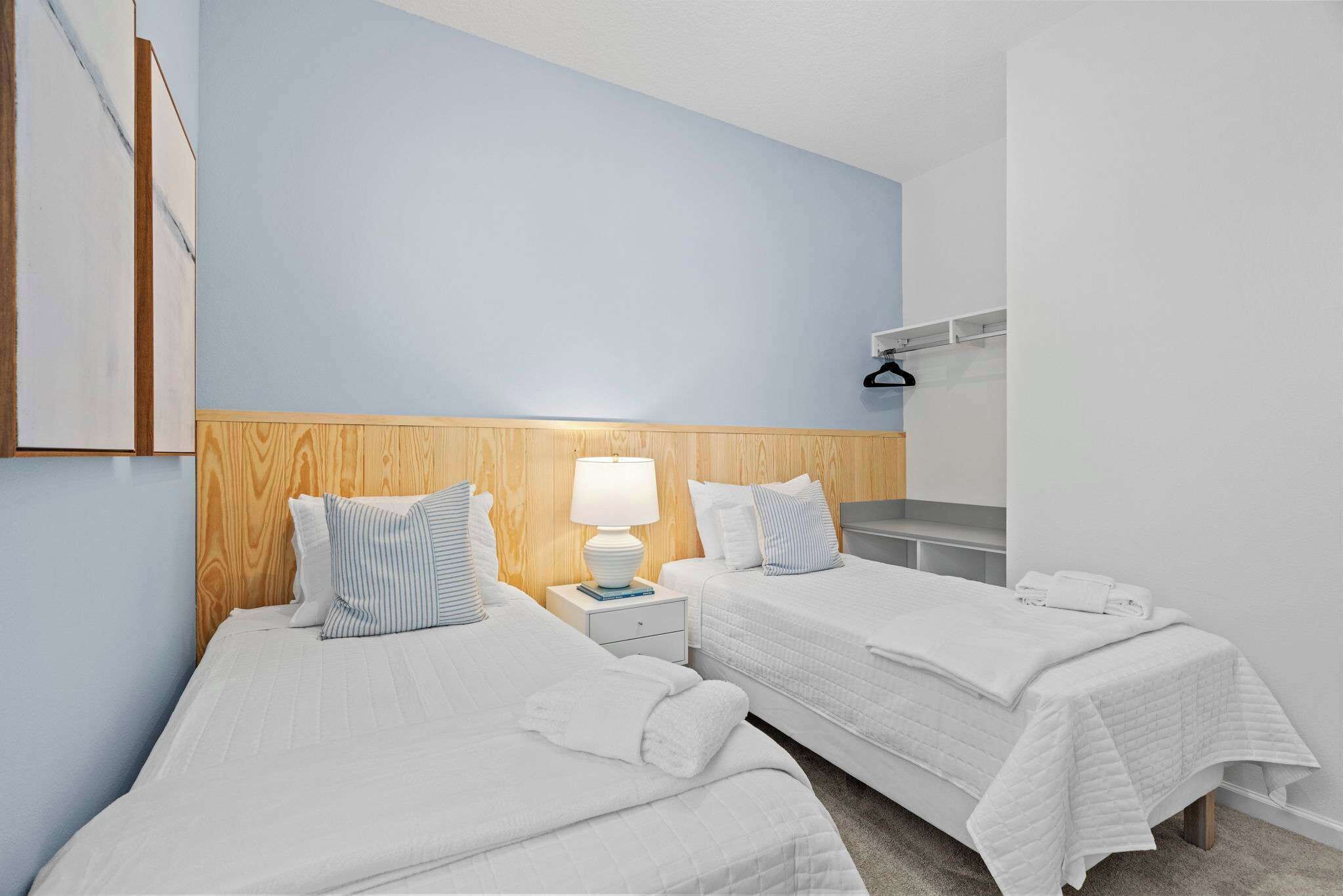 A hotel room with two beds, a wooden headboard, a nightstand with a lamp, and a wall-mounted coat rack.