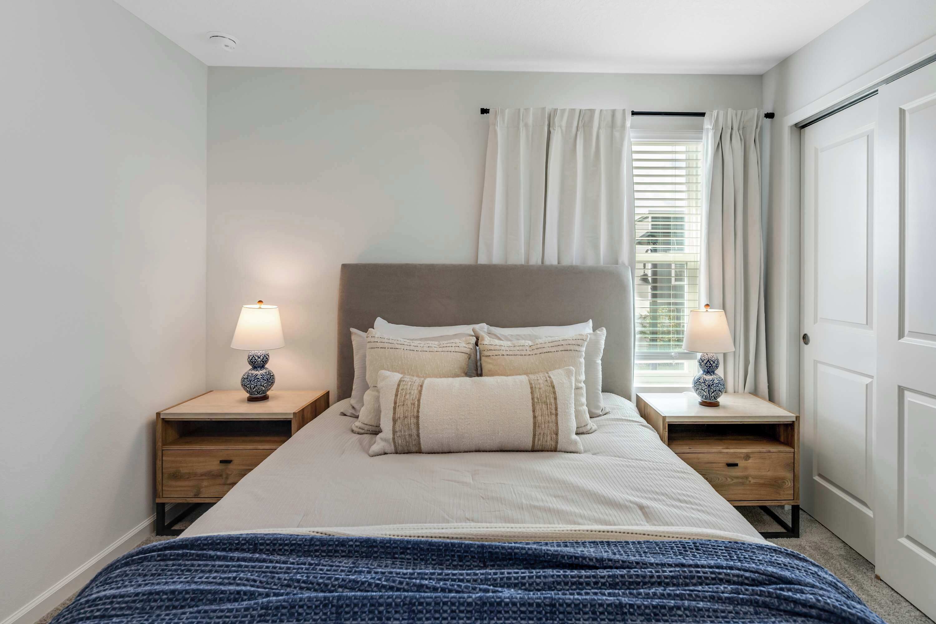 A bedroom with a bed, two bedside tables with lamps, a window with curtains, and a door.