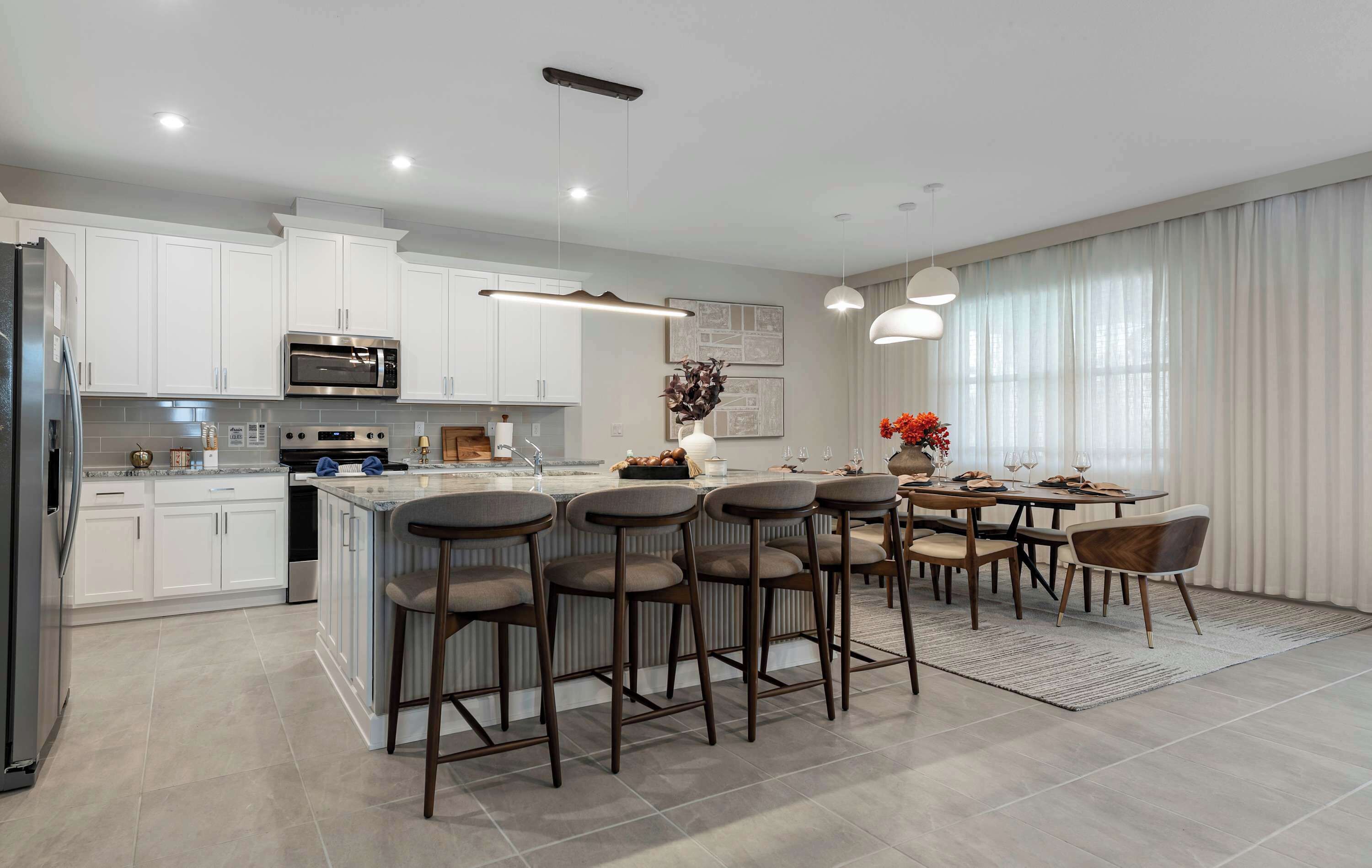 A modern kitchen with white cabinets, a central island with bar stools, a dining area with a table and chairs, and a large window with curtains.