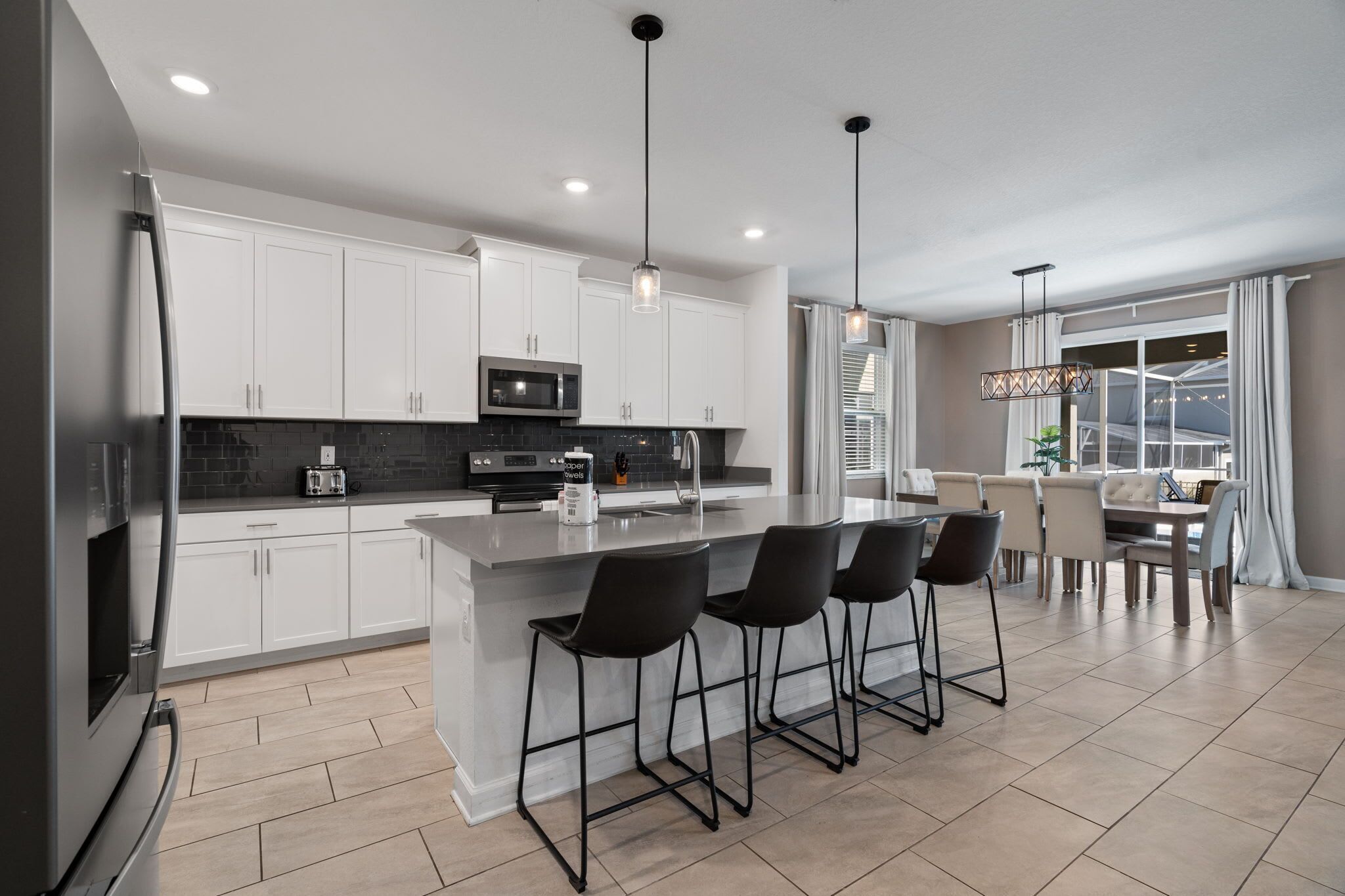 A modern kitchen with white cabinets, a central island with bar stools, a dining area with a table and chairs, and pendant lighting.