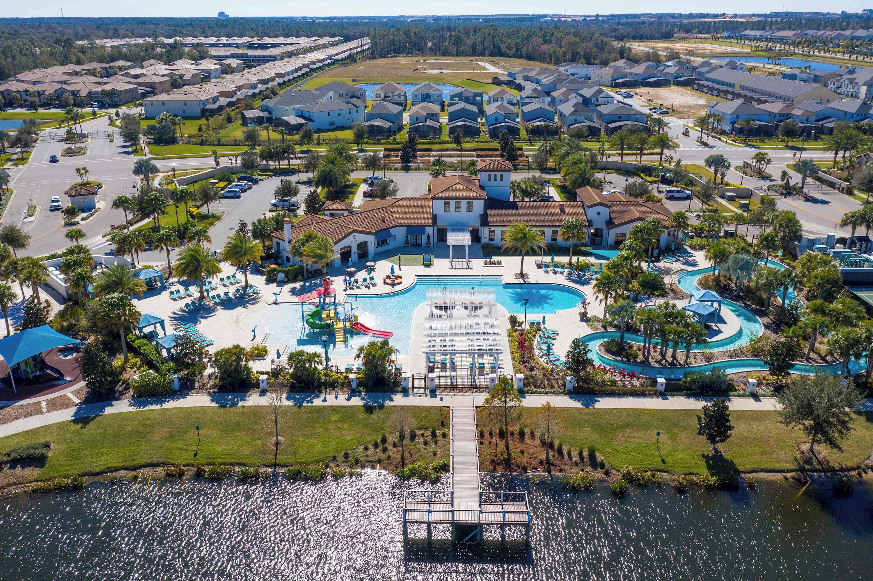 An aerial view of a residential area with a large swimming pool, multiple houses, and a dock extending into a lake.