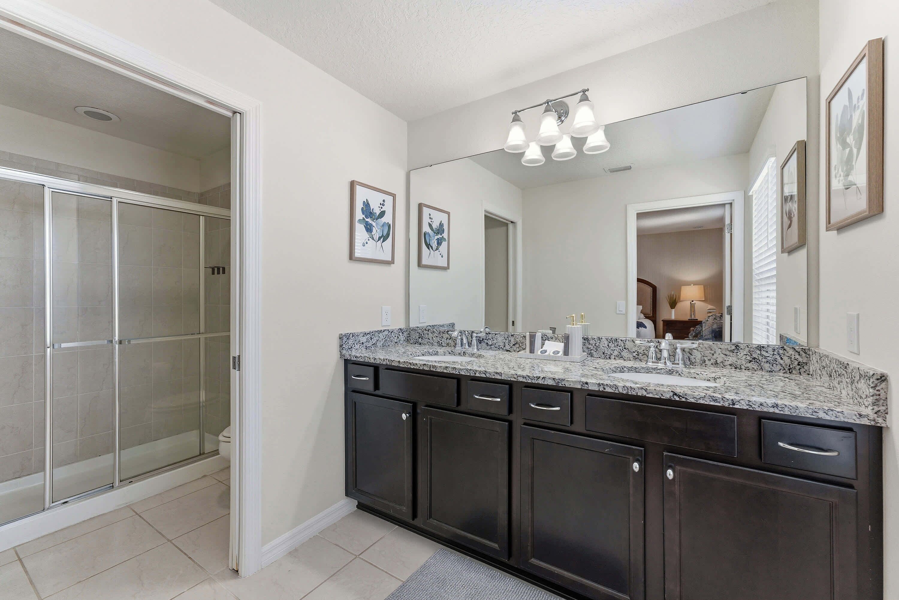A bathroom with a glass shower enclosure, dark cabinetry, a granite countertop, and a large mirror with a light fixture above it.