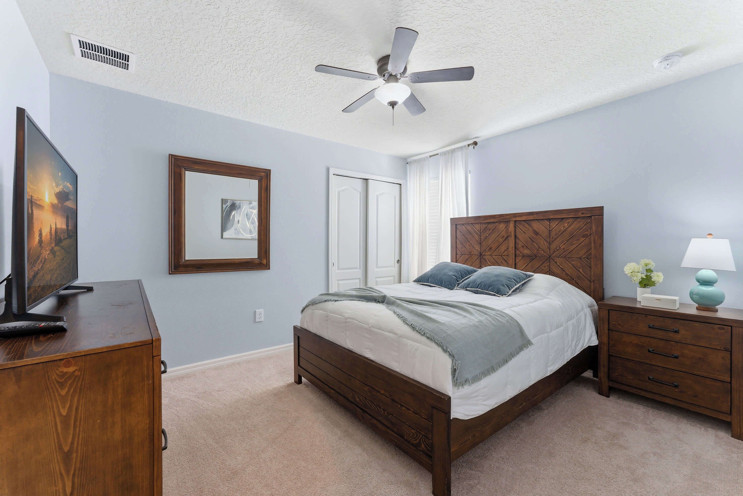 Une chambre à coucher avec un lit, une table de chevet, un bureau, un ventilateur de plafond et un tableau accroché au mur.