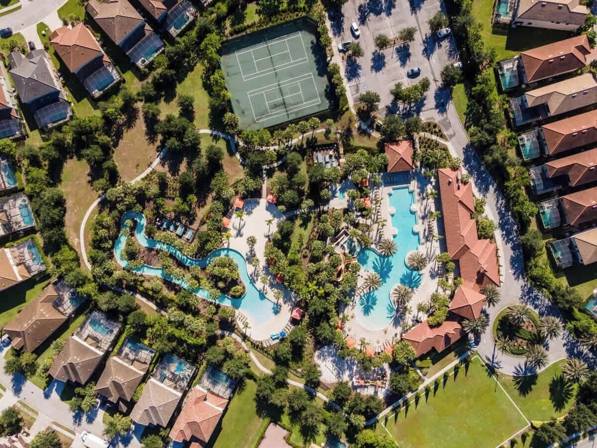 An aerial view of a residential area with a central swimming pool, tennis courts, and a winding water slide.