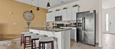 Una cocina moderna con armarios blancos, una isla central con taburetes, un refrigerador grande e iluminación colgante.