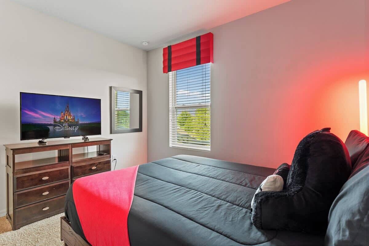 A bedroom with a bed, a TV, a dresser, a window with blinds, and a red curtain.