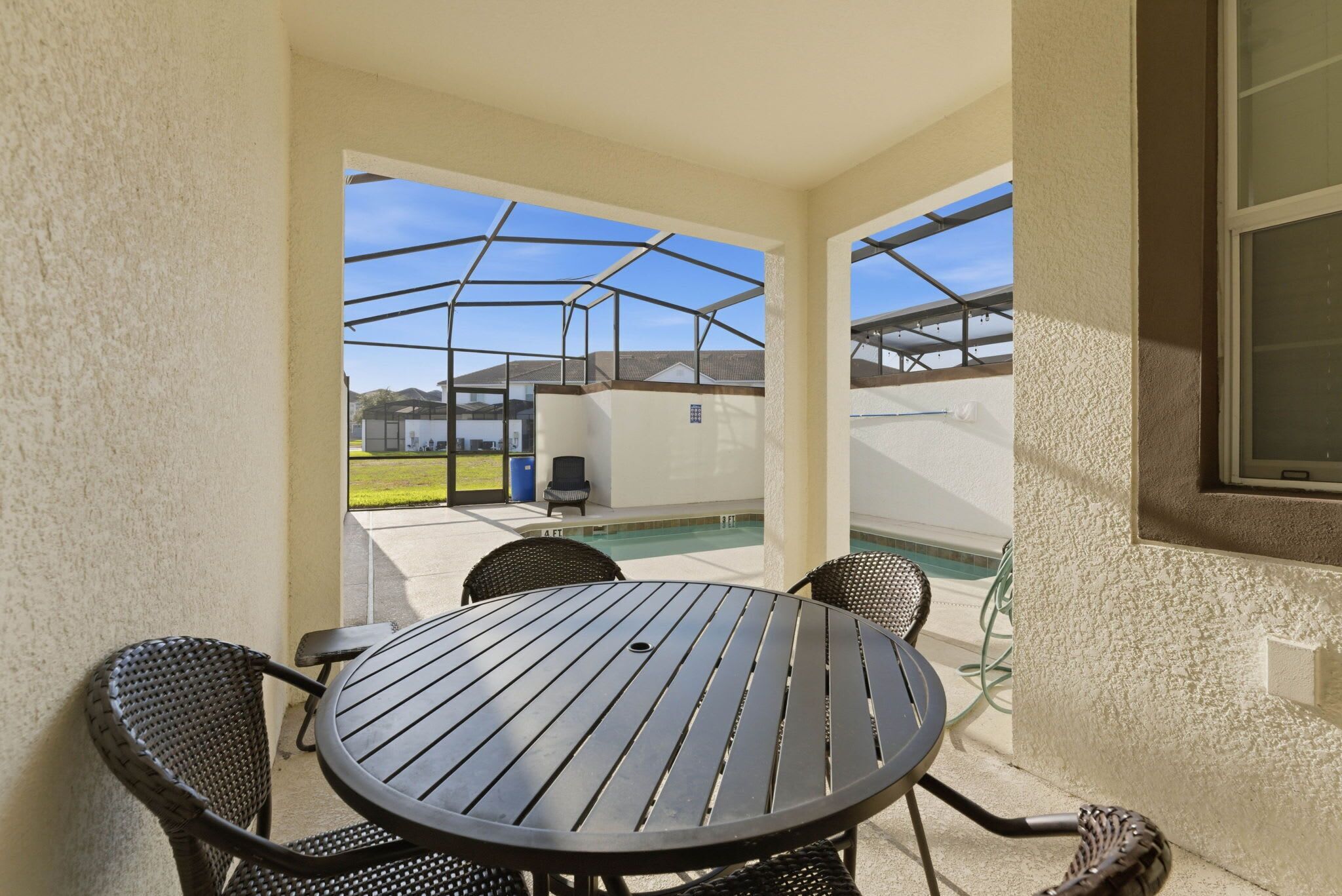 A covered patio area with a round table and chairs, a pool in the background, and a view of neighboring houses.