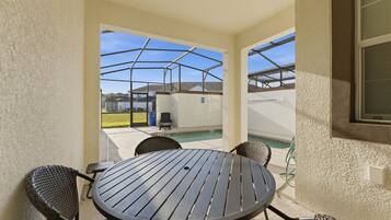 A covered patio area with a round table and chairs, a pool in the background, and a view of neighboring houses.