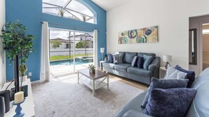 Ein modernes Wohnzimmer mit blauer Akzentwand, einem grauen Sofa mit blauen Kissen, einem Couchtisch und einem großen Fenster mit Blick auf einen Pool.