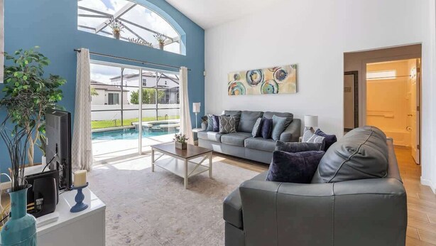 Ein modernes Wohnzimmer mit grauem Sofa, einem Couchtisch und einem Fernseher. Ein großes Fenster bietet Blick auf einen Swimmingpool und eine blaue Wand.