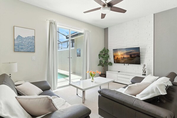 A modern living room with a flat-screen TV, a ceiling fan, and a large window with curtains. - Relax Near Disney, 4BR Home With Pool (Kissimmee)