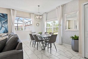 A dining area with a glass table, grey chairs, and a chandelier. There is a grey sofa in the foreground and a potted plant near the door. - Disney Familly Lovers up to 11 Guests (Kissimmee)