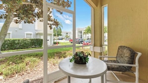 A balcony with a white table and chair, a potted plant, and a view of a residential area with buildings and parked cars. - Resort Condo 3BR - Pool, Balcony, Near Disney (Kissimmee)