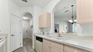 A modern kitchen with white cabinetry, a dishwasher, a countertop with a sink, and a large mirror above the sink. - Resort Condo 3BR - Pool, Balcony, Near Disney (Kissimmee)