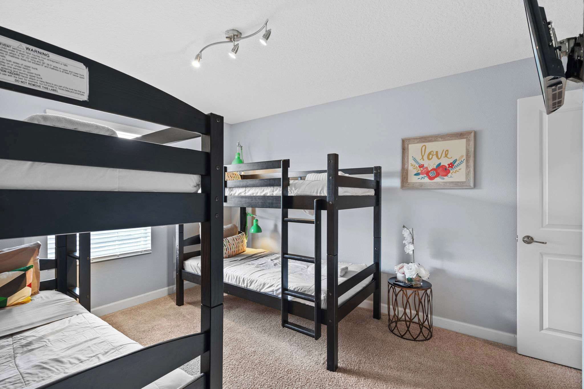 A bunk bed room with a warning label, a small table with a lamp, and a framed picture on the wall.