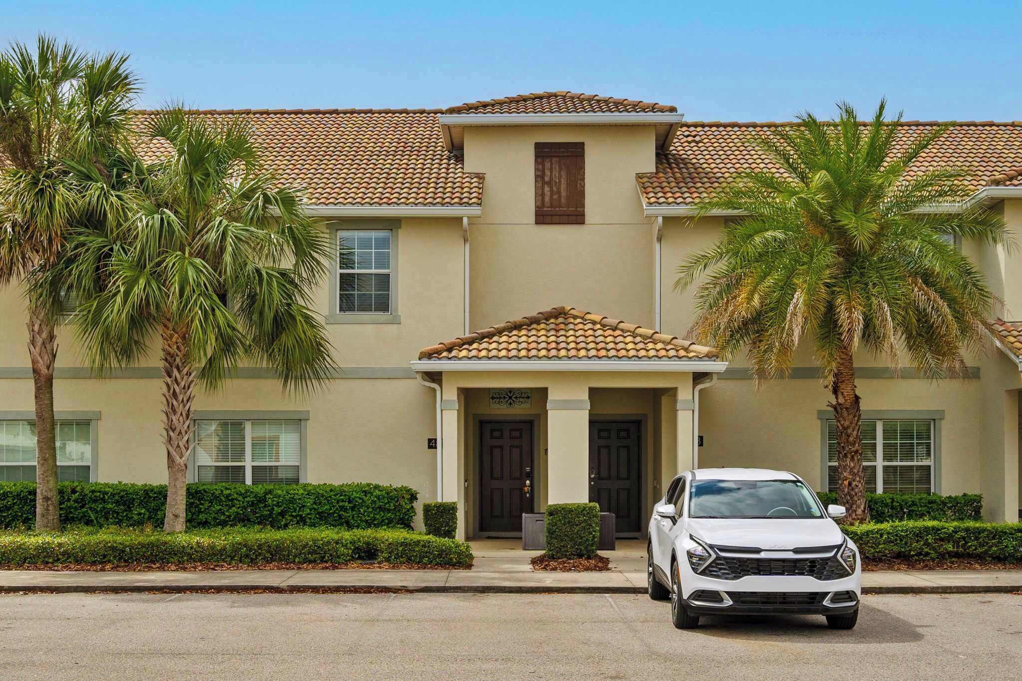 A two-story building with a tiled roof, palm trees, and a white car parked in front.