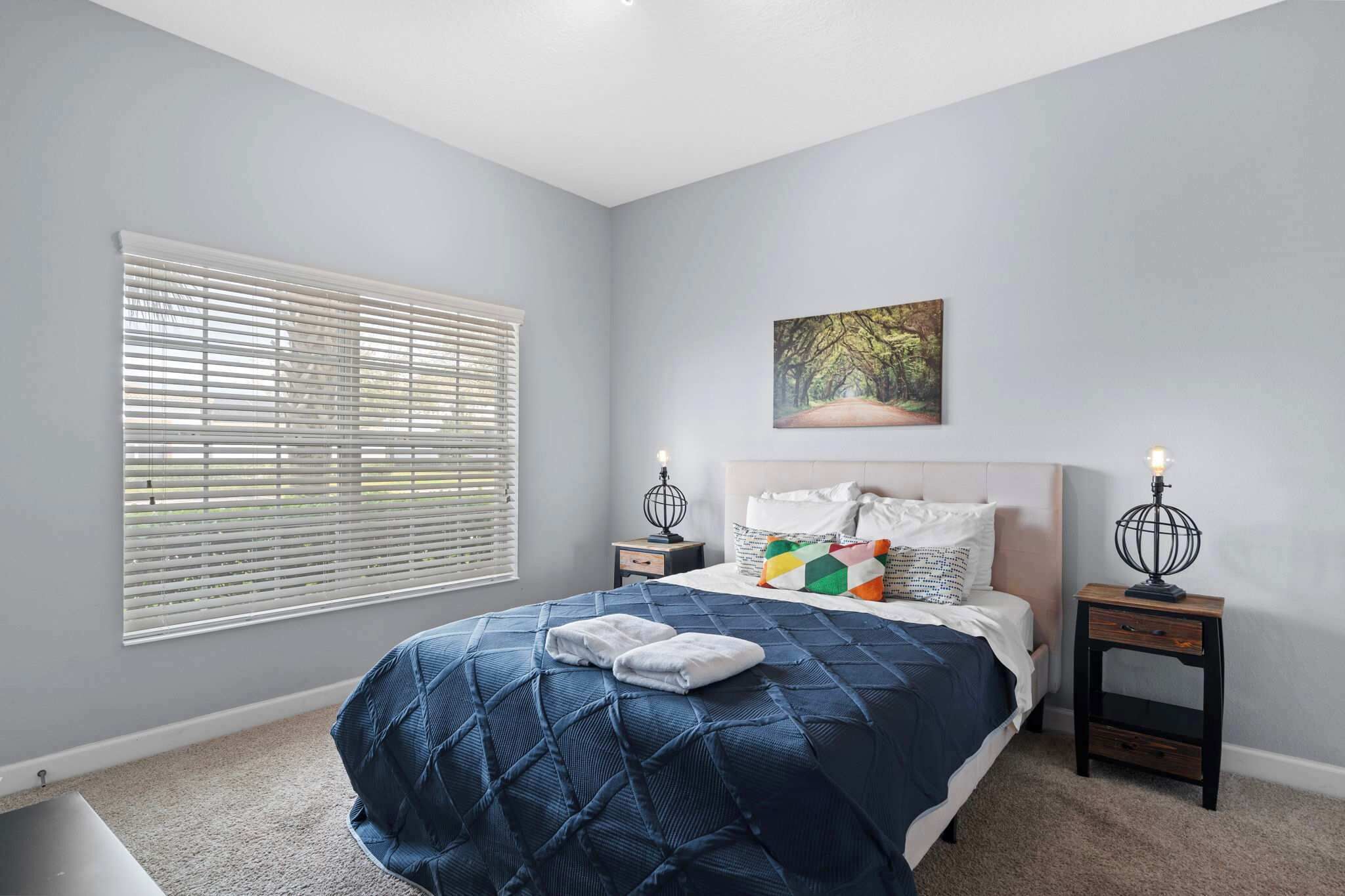 A bedroom with a bed, two bedside tables, a nightstand, a window with blinds, and a painting on the wall.