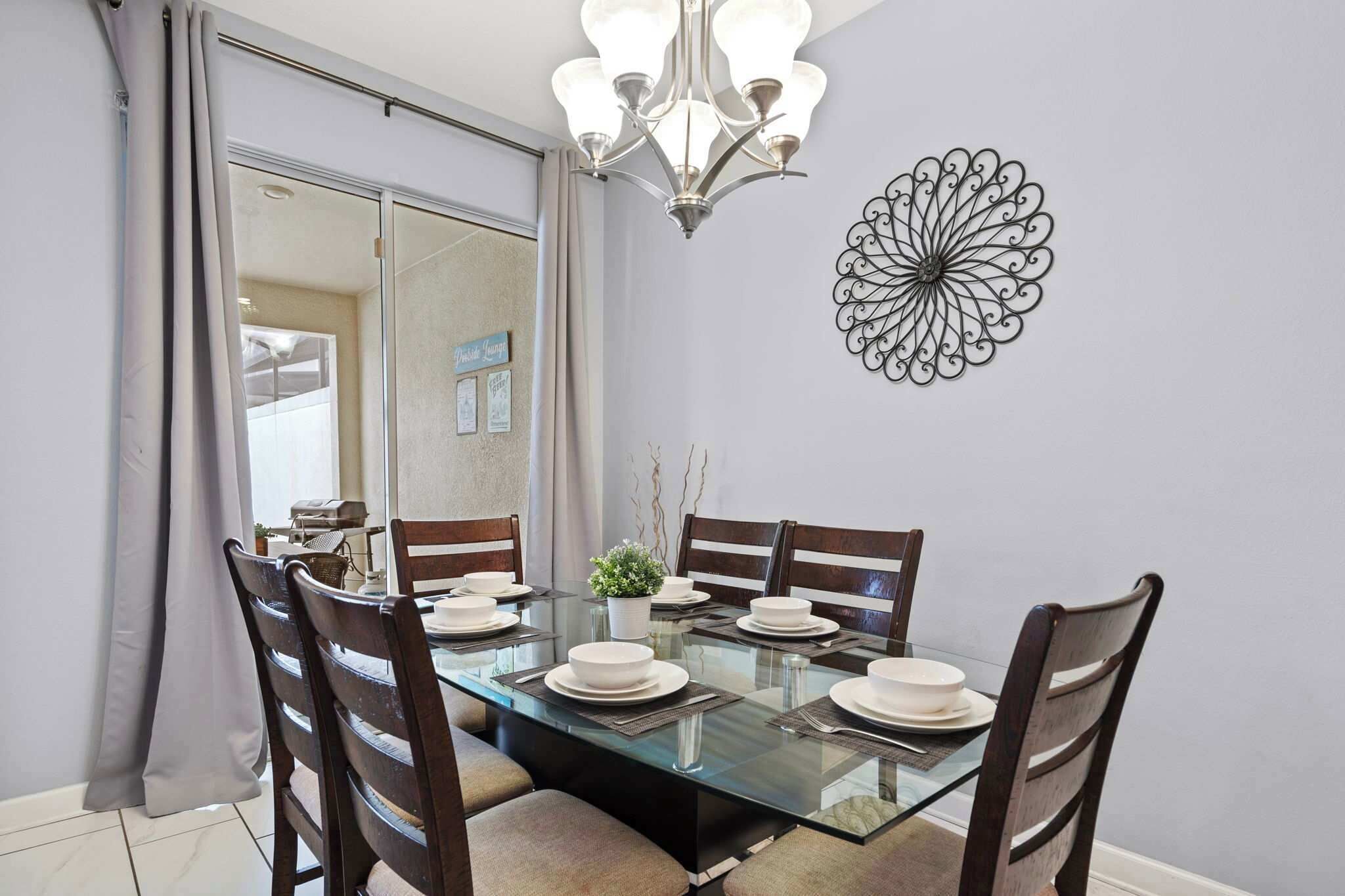 A dining area with a glass table, wooden chairs, a chandelier, and a decorative metal wall piece.