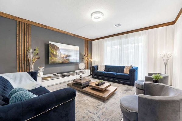A modern living room with a dark blue sofa, a wooden coffee table, and a grey armchair. A large painting is on the wall, and there is a white console table with decorative items. - Sophisticated 6BR at Storey Lake (Kissimmee)