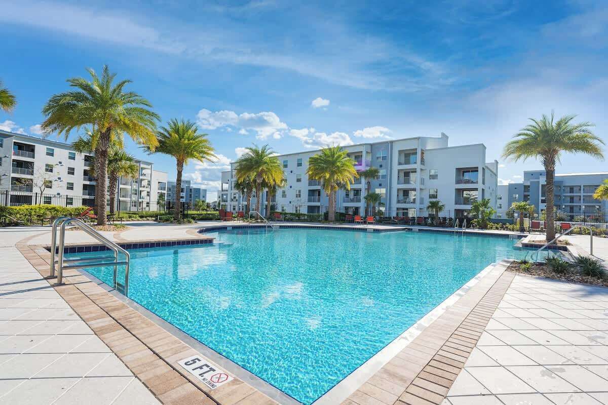 A swimming pool surrounded by modern buildings, palm trees, and a clear sky.