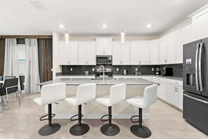 A modern kitchen with white cabinets, a central island with bar stools, a refrigerator, and a microwave. - Sophisticated 6BR at Storey Lake (Kissimmee)