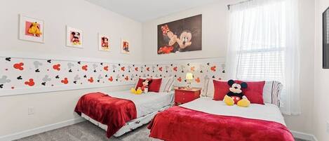 Une chambre avec deux lits, chacun agrémenté de linge de lit aux motifs de Mickey Mouse, un doudou à l’effigie de Mickey Mouse sur le plus grand lit, et des décorations murales représentant Mickey Mouse.