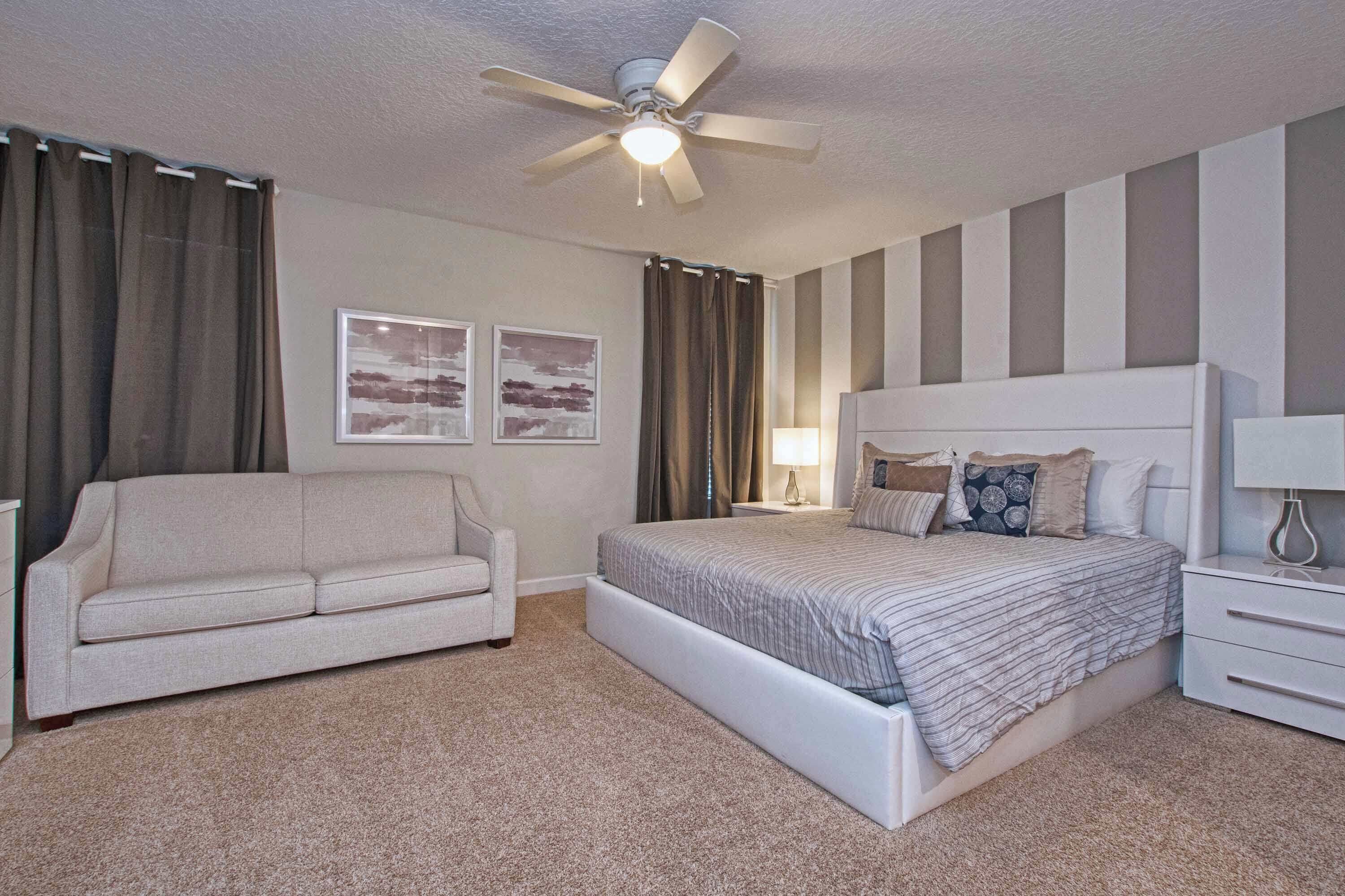 A bedroom with a bed, a nightstand, a lamp, a ceiling fan, a couch, and two framed pictures on the wall.