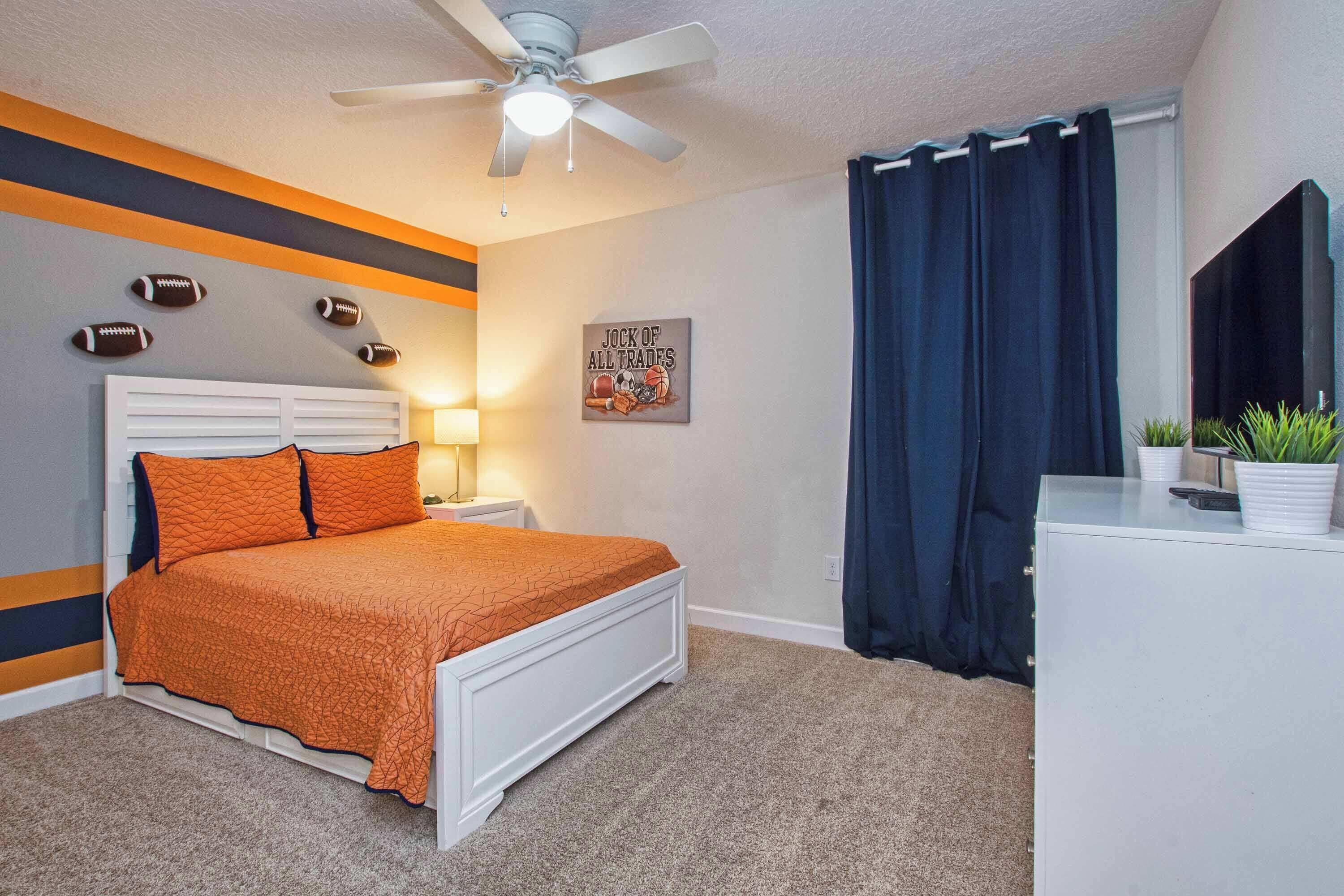 A bedroom with a bed, a ceiling fan, a nightstand, a dresser, a TV, and a wall with football decorations.