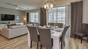 A modern hotel room with a dining area, a sofa, a flat-screen TV, and a large window with curtains. - Spacious 6 Bed Home With Pool in Storey Lake (Kissimmee)