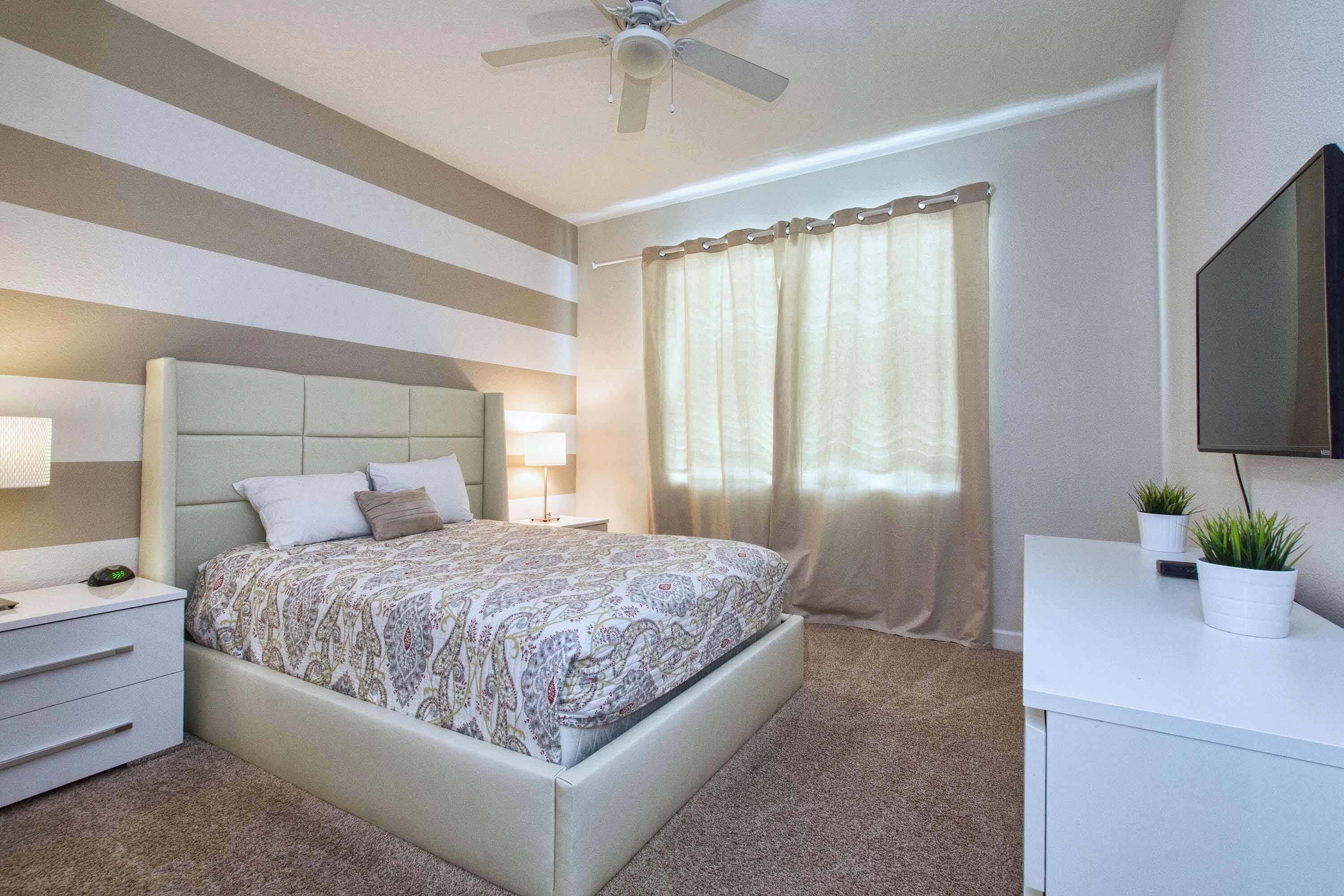 A bedroom with a bed, bedside tables, a ceiling fan, a TV, and curtains.