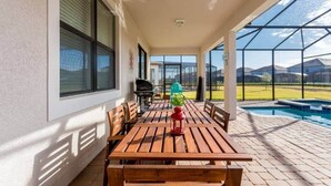 Een overdekt terras met een houten tafel en stoelen, een barbecue en een zwembad op de achtergrond.