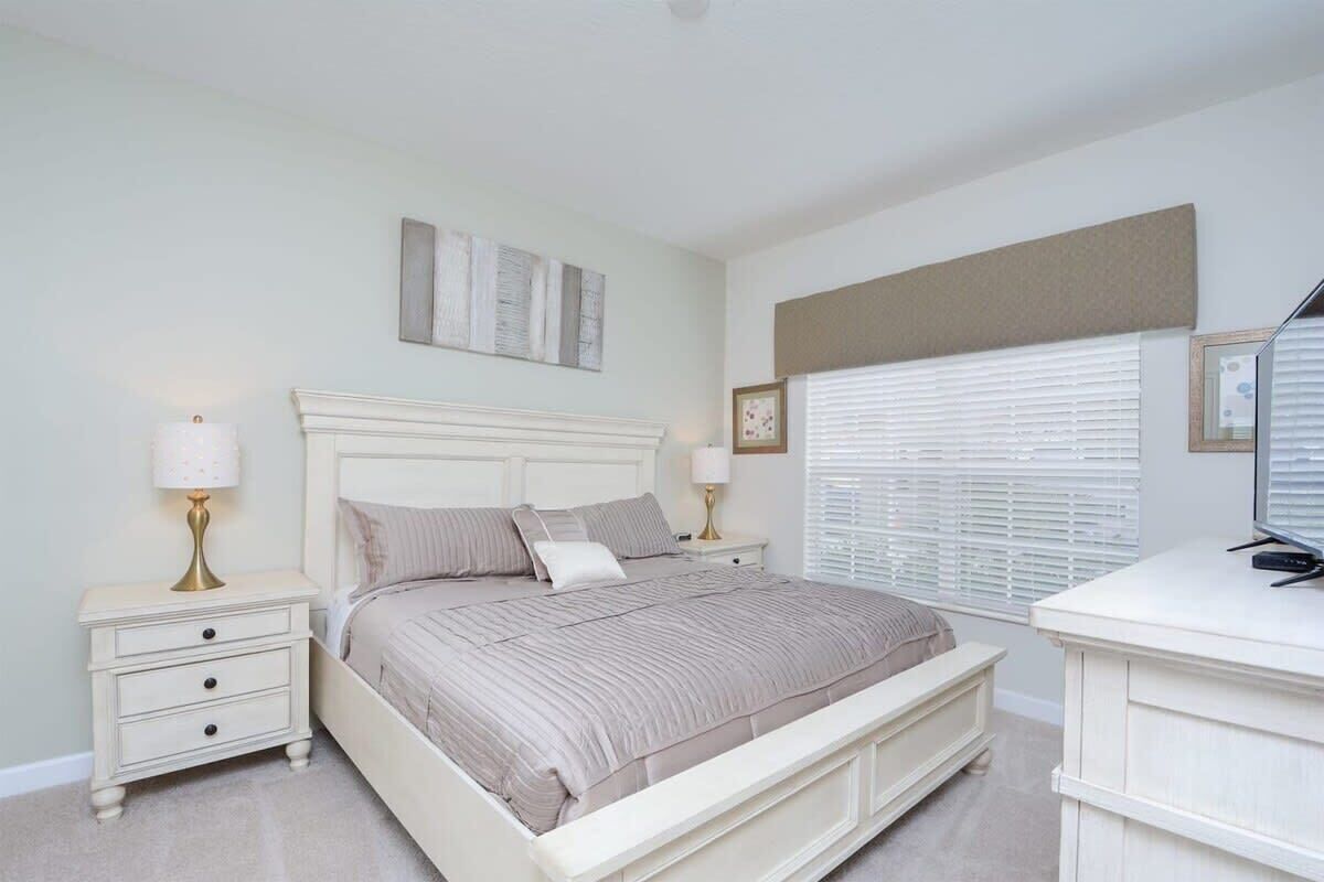 Une chambre à coucher comprenant un lit, des tables de chevet, un bureau et une fenêtre avec des stores.