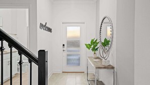 Un couloir avec une pancarte « Bienvenue », une porte en verre dépoli, une plante en pot, un miroir orné de perles et un escalier avec des balustres noires.