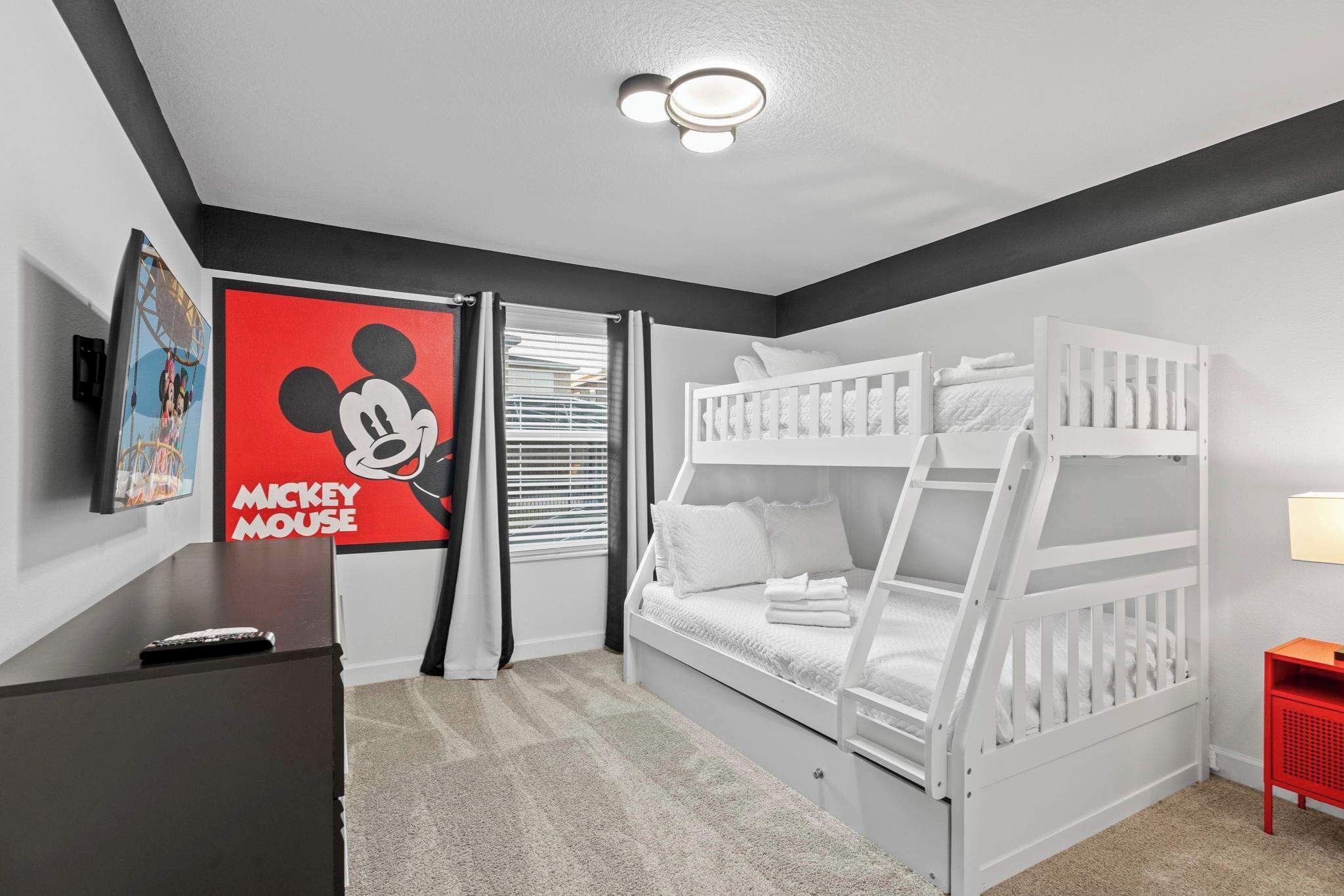 A room with bunk beds, a Mickey Mouse poster, a desk with a remote control, and a red side table.
