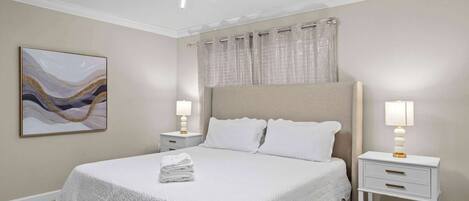 A neatly made bed with a white bedspread and pillows, two bedside tables with lamps, a ceiling light fixture, and a wall-mounted abstract painting.