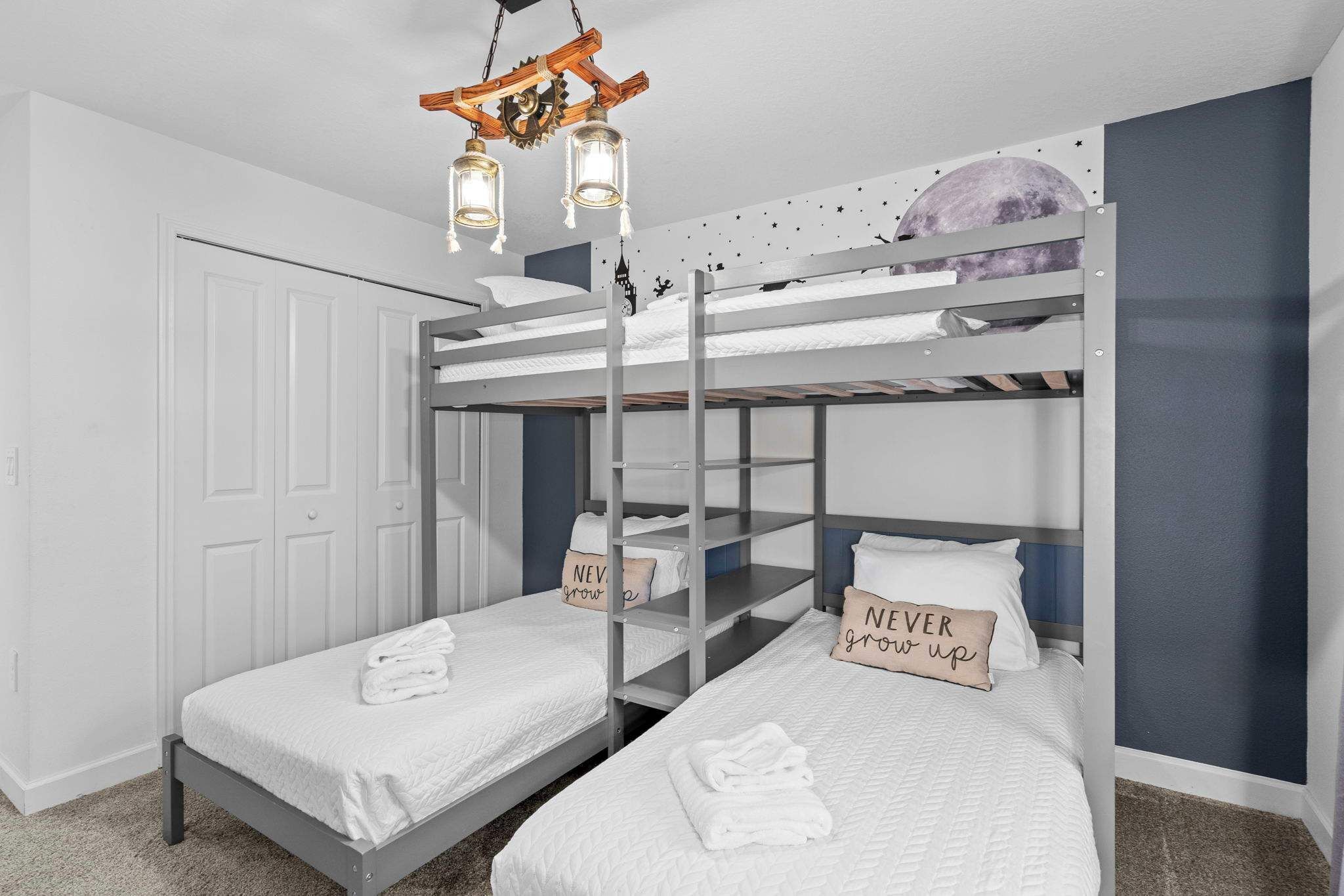 Une chambre avec un lit superposé, du linge de lit blanc, un luminaire en bois au plafond et un autocollant représentant la lune sur le mur.