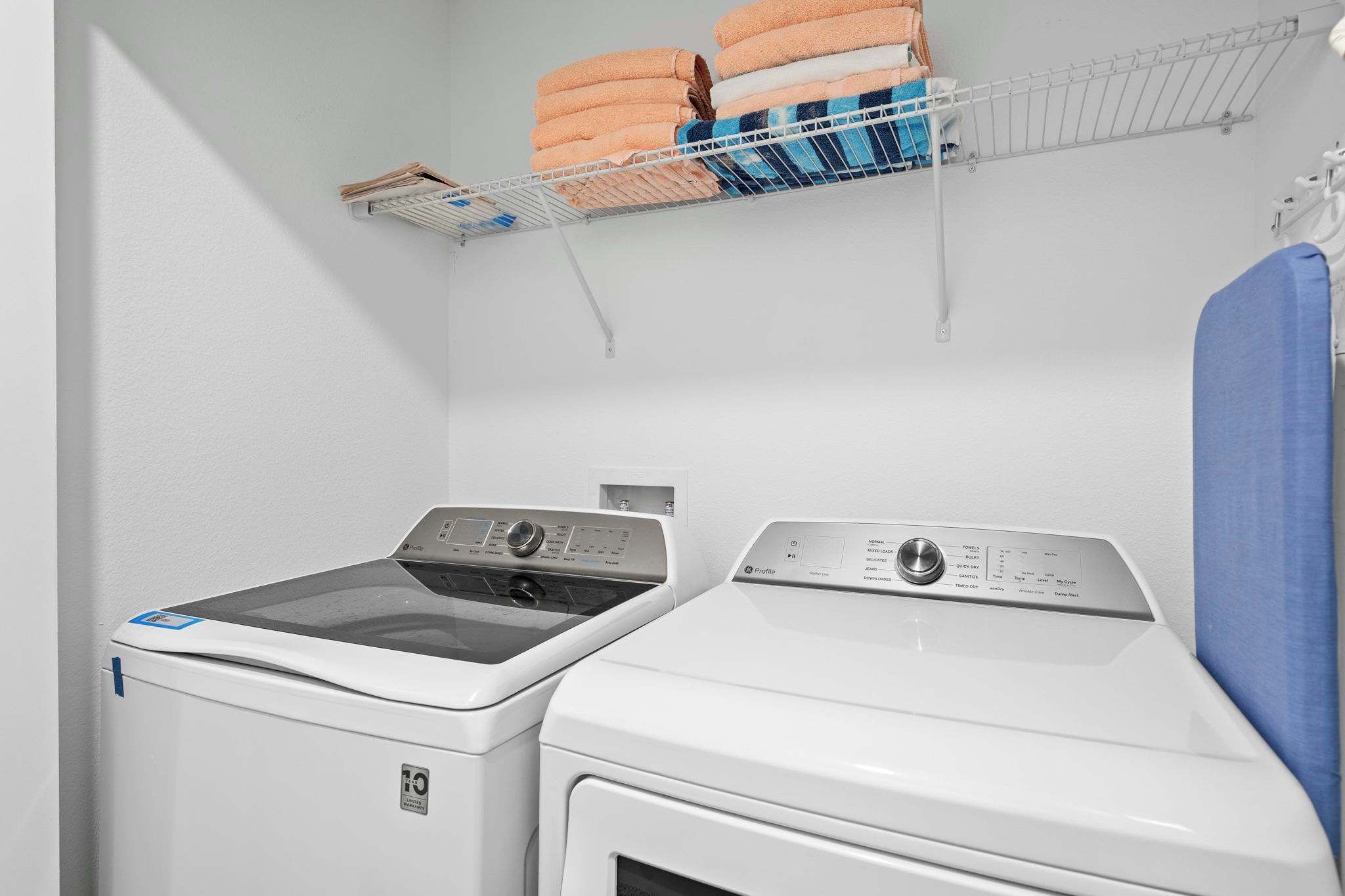 Une buanderie équipée de deux machines à laver à chargement frontal, une étagère en métal au-dessus où sont pliées des serviettes, et une serviette bleue suspendue sur le côté droit.