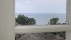 Vista costera a través de una ventana, con un mar tranquilo, cielo parcialmente nublado y edificios tradicionales con techos de paja.