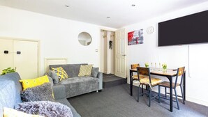 Un salon comprenant un canapé, une table à manger avec des chaises, un téléviseur à écran plat fixé au mur et une porte donnant sur une autre pièce.