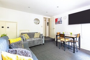 Ein Wohnzimmer mit einer Couch, einem Esstisch mit Stühlen, einem flachbildfernseher an der Wand und einer Tür zu einem anderen Raum.
